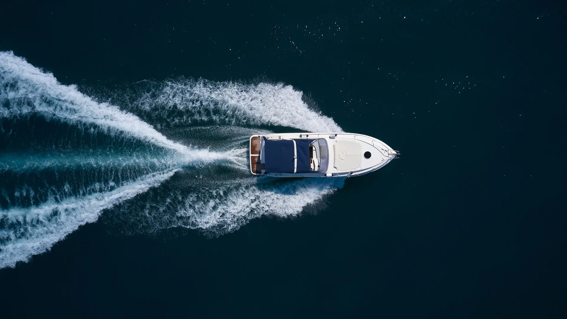 Aerial view of a white motorboat cruising on deep blue water, leaving two curved trails of white wake behind.   Image depicting the Marine market