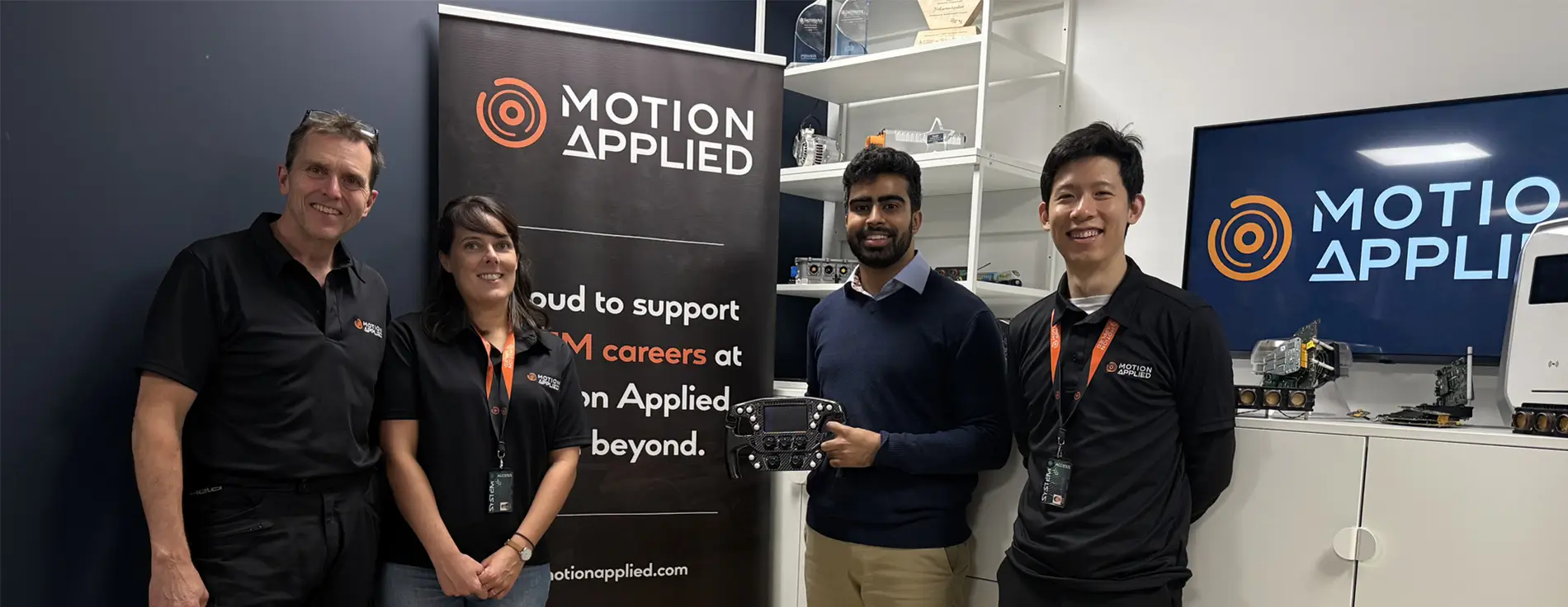 Group of people standing in a Motion Applied office or lab, with Motion Applied branding visible on banners and a screen, and one person holding an electronic control unit in front of display shelves